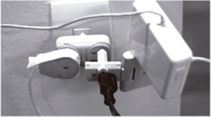Fotografia em preto e branco de um adaptador de tomadas (conhecido popularmente como 'benjamim' ou 'T') conectado à parede, com múltiplos plugues de diferentes aparelhos inseridos nele simultaneamente.