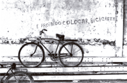 Fotografia em preto e branco mostrando uma bicicleta estacionada e encostada em um muro. No muro, logo acima da bicicleta, há uma inscrição pintada à mão que diz: 'É PROIBIDO COLOCAR BICICRETA'.